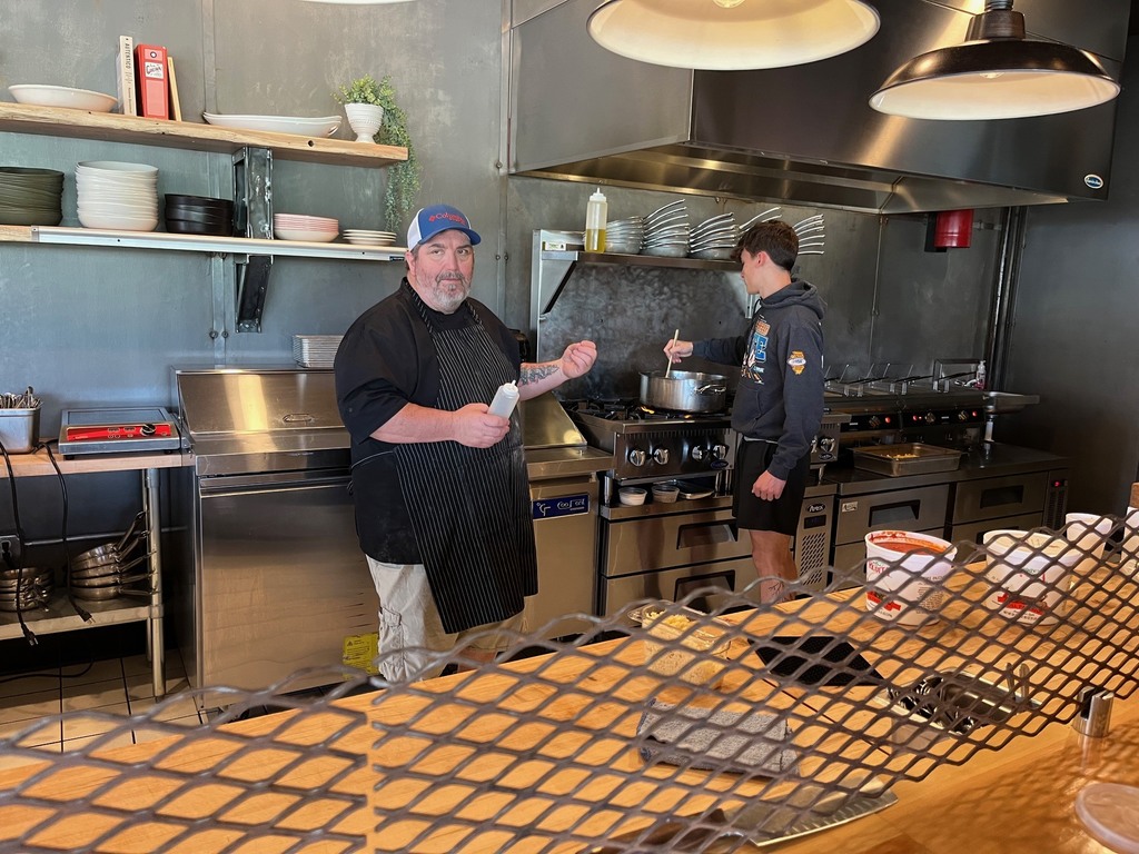 The chef is talking and a student is stirring something in a pot on the stove. 