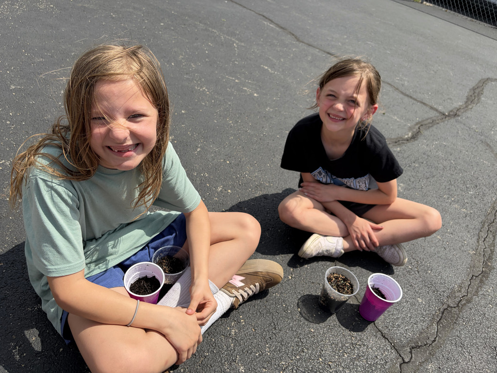 Two students are sitting on the blacktop with their plant cups. 