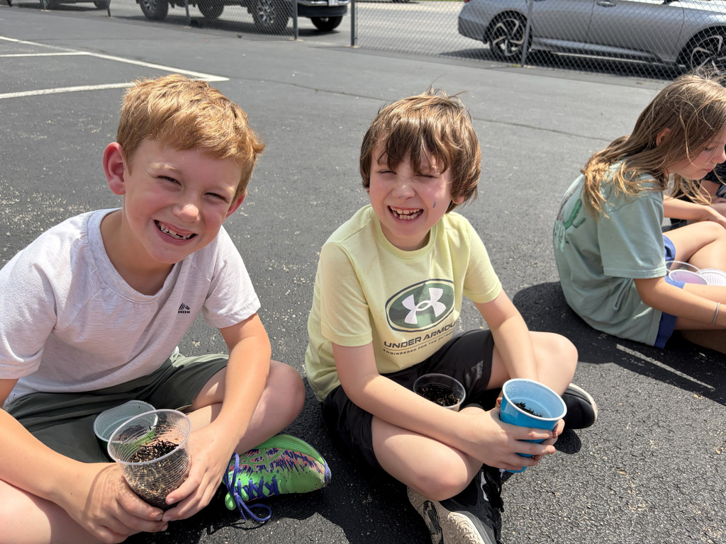 Two students are sitting on the blacktop with their plant cups. 