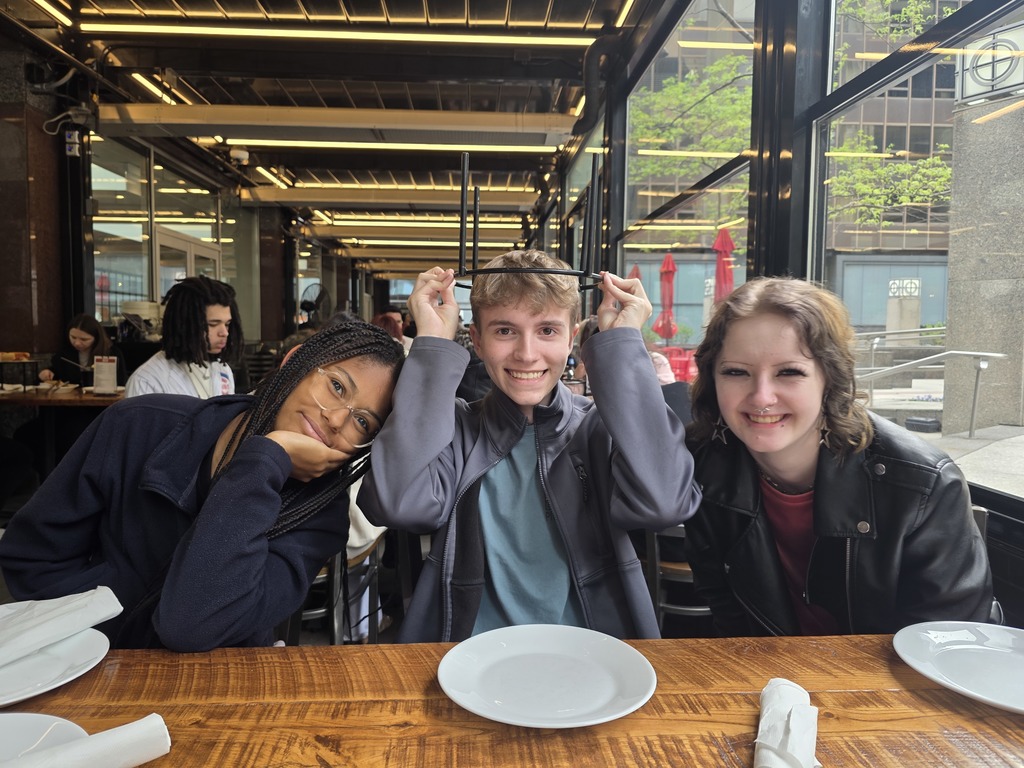 Three students are sitting at a table. One is holding the metal pizza holder over his head like a hat. 
