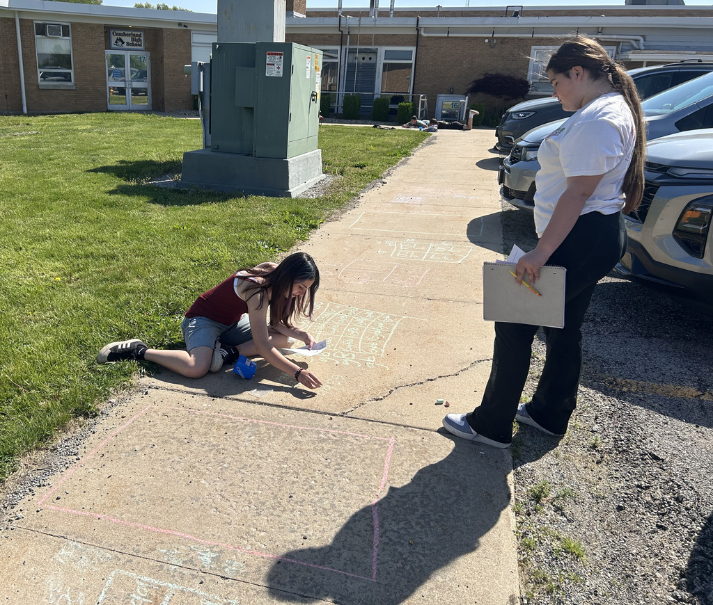 A student is working on a Punnett square with sidewalk chalk with the help of another student. 