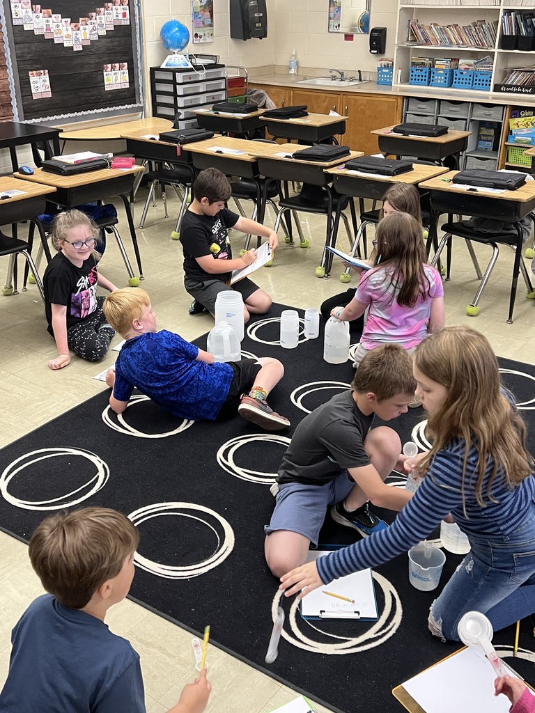 Seven students are sitting on the carpet. They are measuring different liquids and making notes on their clipboards. 