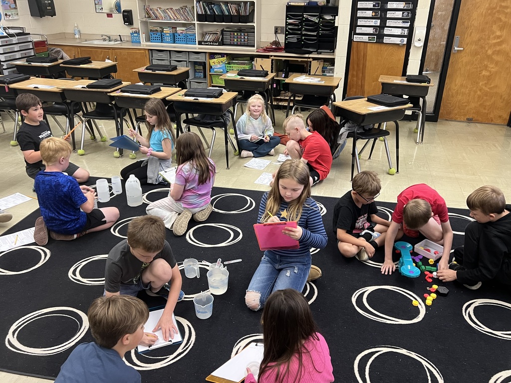 The class is sitting in small groups on the carpet. They are measuring different liquids and making notes on their clipboards. Another group is weighing objecs to compare. 