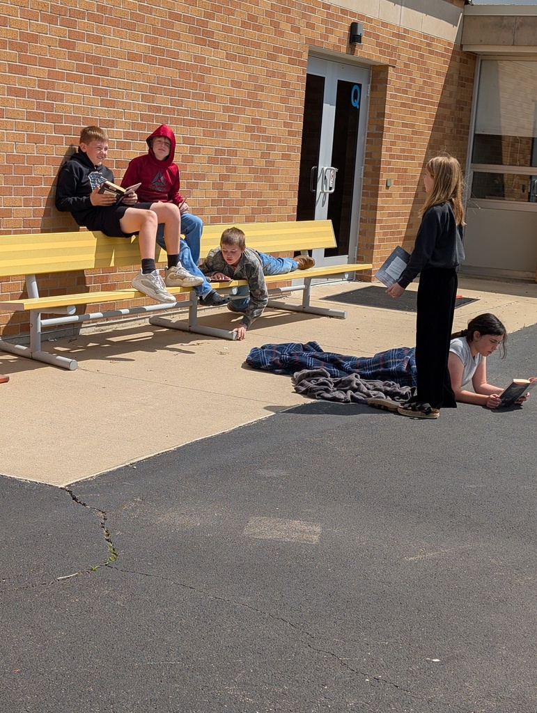 A group of five students are reading their books together at bench and on the blacktop. 