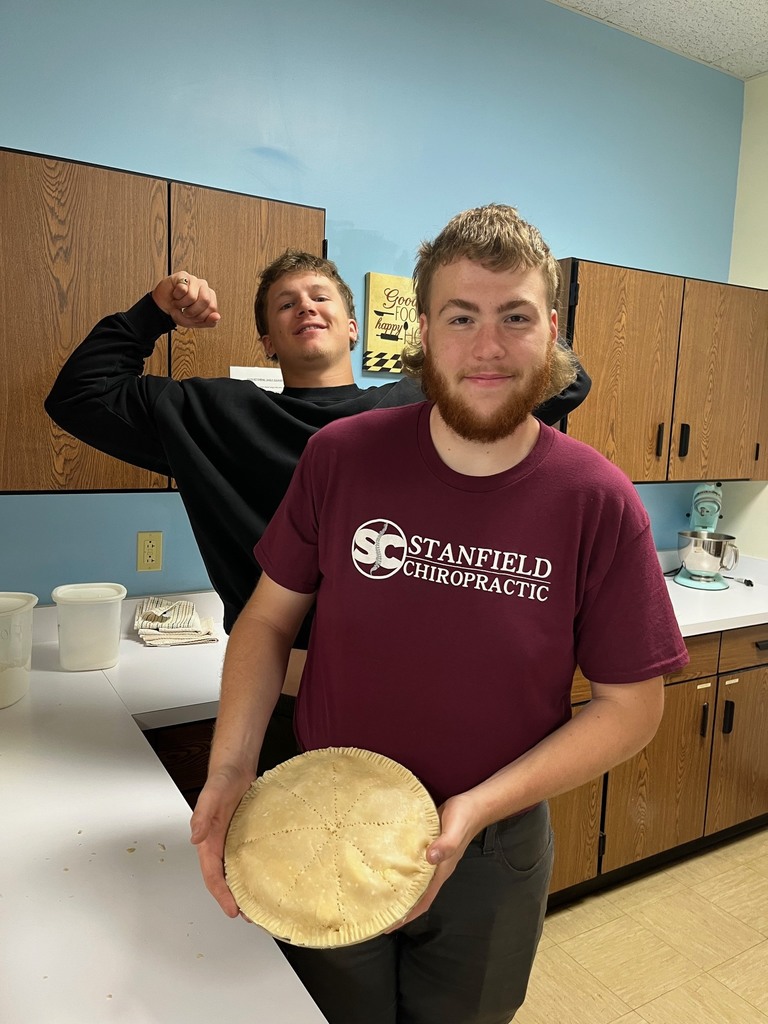 A student is holding a finished apple pie. Another student si flexing in the background. 