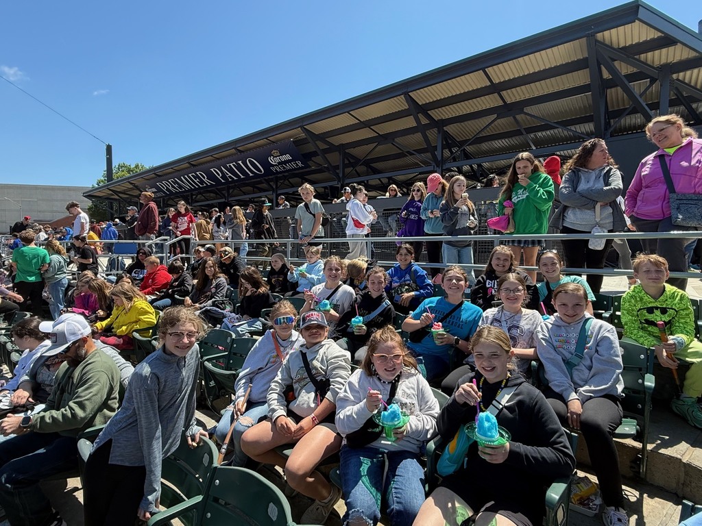 A group of sstudents is sitting in the bleachers watching the baseball game. Many have a souvenier and are eating a snack. 