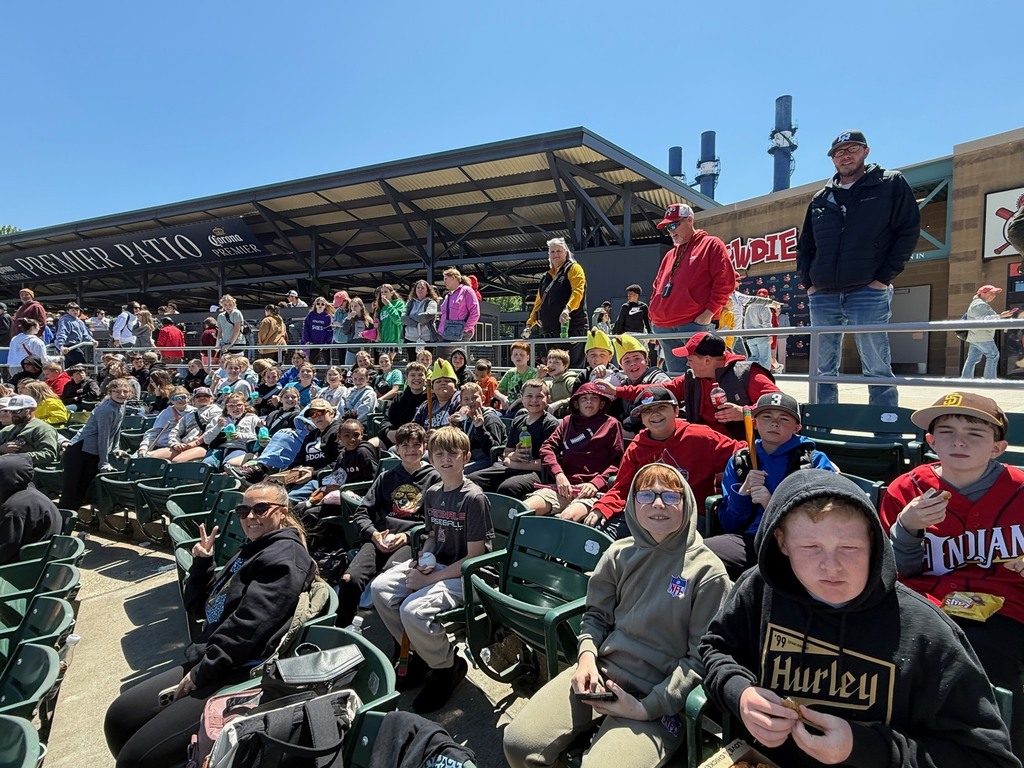 A group of sstudents is sitting in the bleachers watching the baseball game. Many have a souvenier and are eating a snack. 
