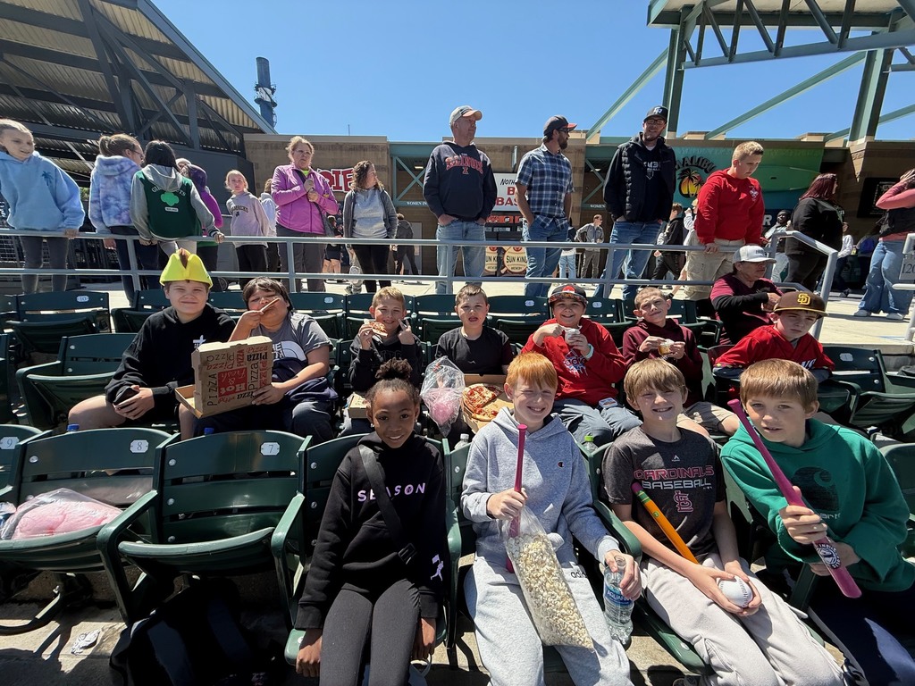 A group of sstudents is sitting in the bleachers watching the baseball game. Many have a souvenier and are eating a snack. 