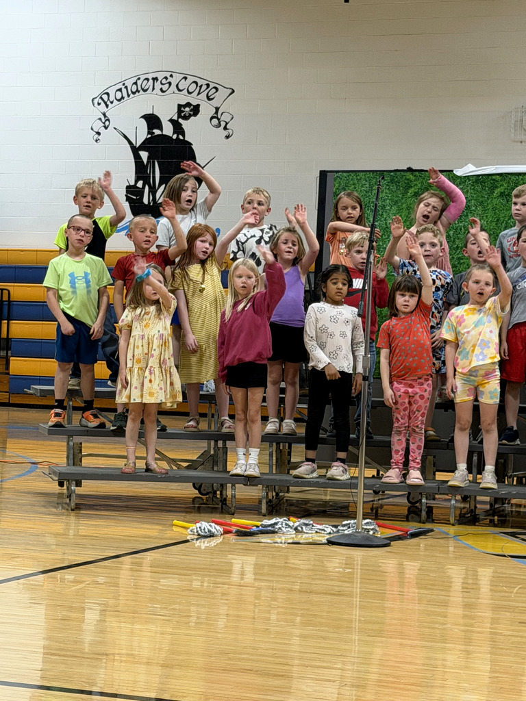 A group of students are standing on risers singing a song and raising their arm.