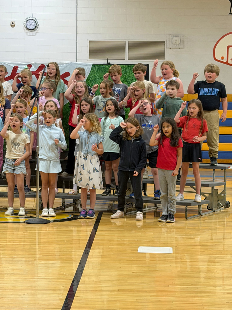 A group of students are standing on risers singing a song and giving the You're out sign.