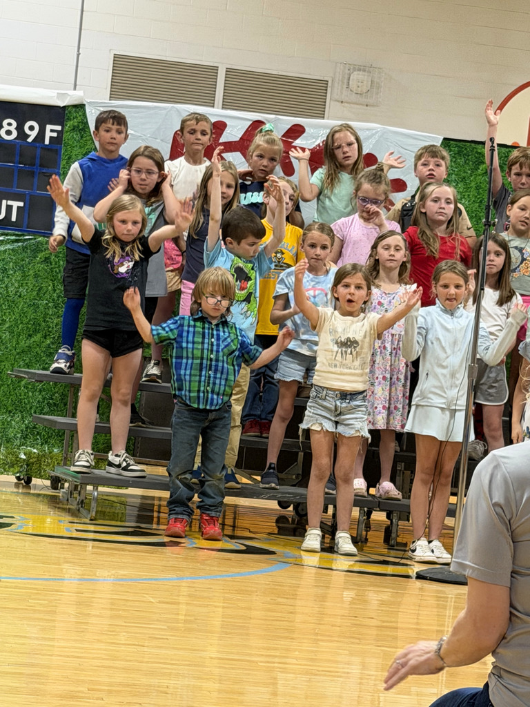 A group of students are standing on risers singing a song and raising their hands. 