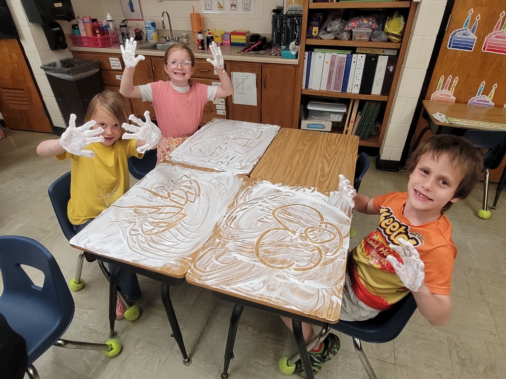 Three students hve shaving cream all over the desk. Their hands are  full, too. 