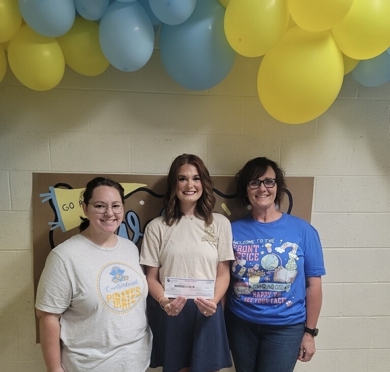 Picture shows three adults. The adult in the middle is holding a check. There are balloons flanking the picture. 