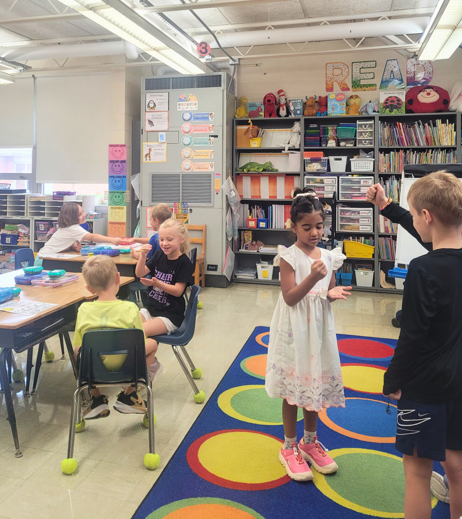 Students are sitting at desks and standing around the room. They are paired and playing rock, paper, scissors. 