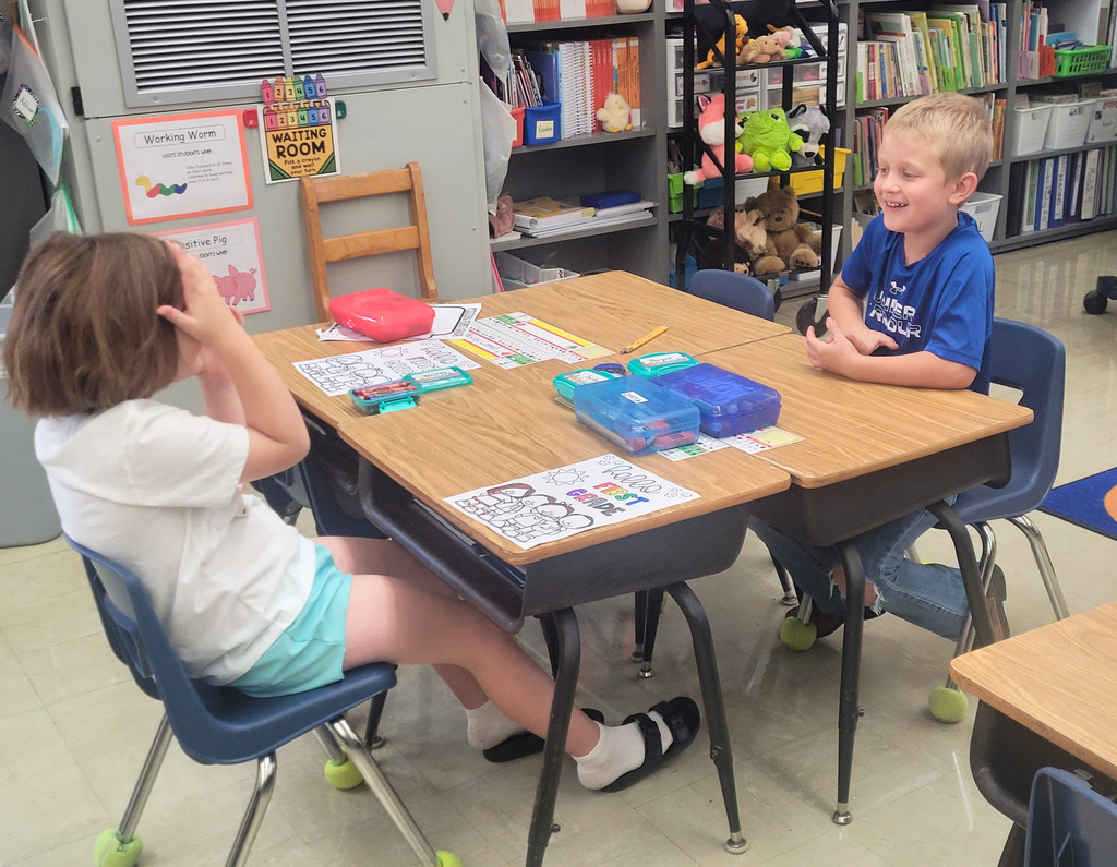 Two students are sitting at their desks. They are playing Rock, Paper, Scissors. One student is covering their face because they lost. 