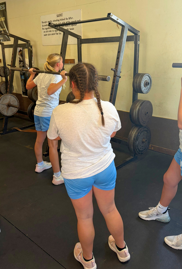 Pictured is a student with a bar on their shoulders preparing to do squats. Another student is watching on. 