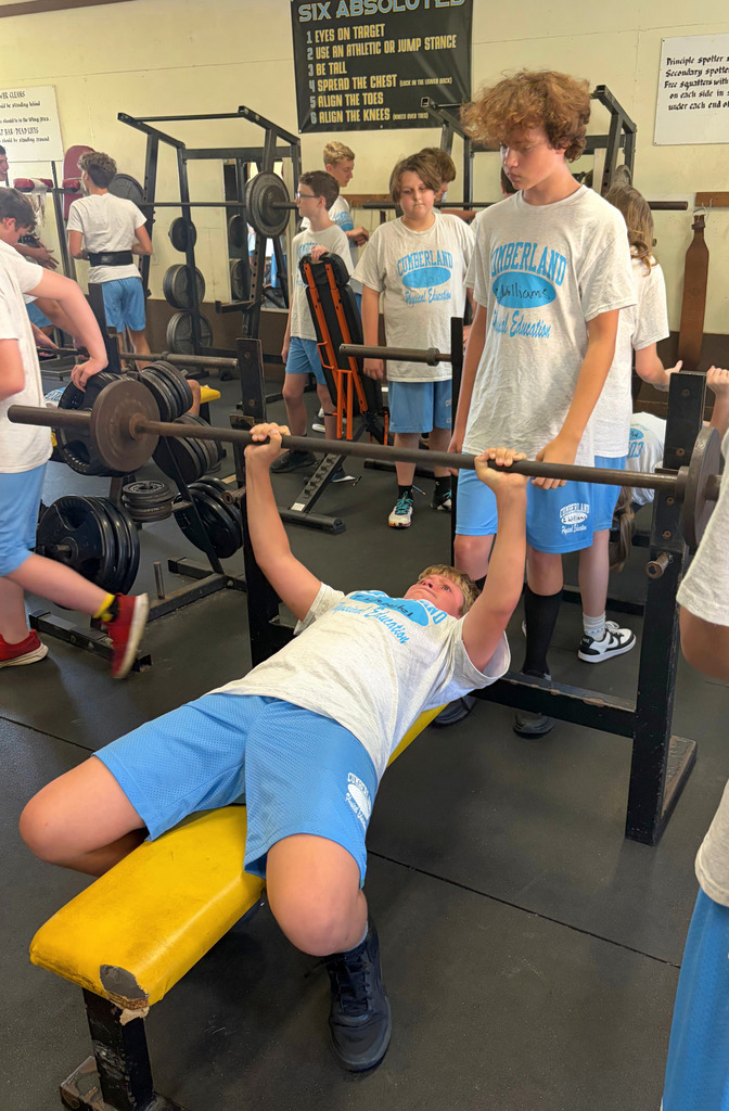 Picured is a student bench pressing. Another student is spotting. Other students are at different parts of the weight room doing other activities. 