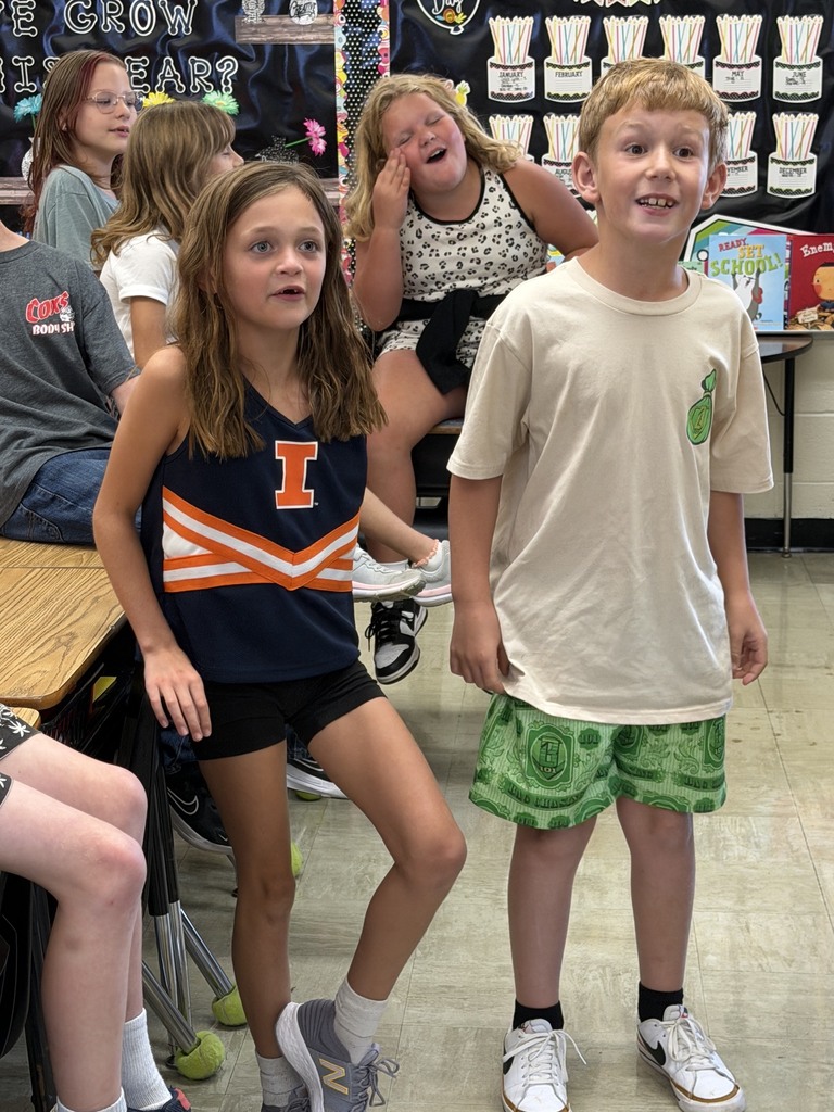 Two students are standing side by side answering a multiplication problem as fast as they can. Other students in the class are watching. 