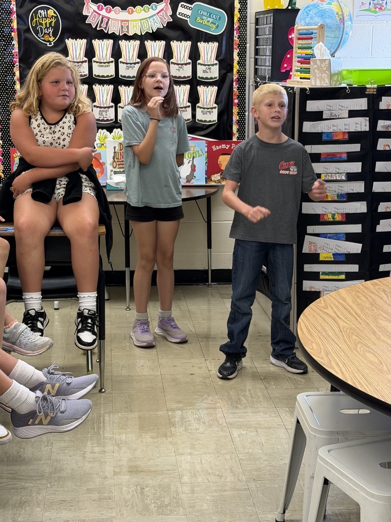 Two students are standing side by side answering a multiplication problem as fast as they can. Other students in the class are watching. 