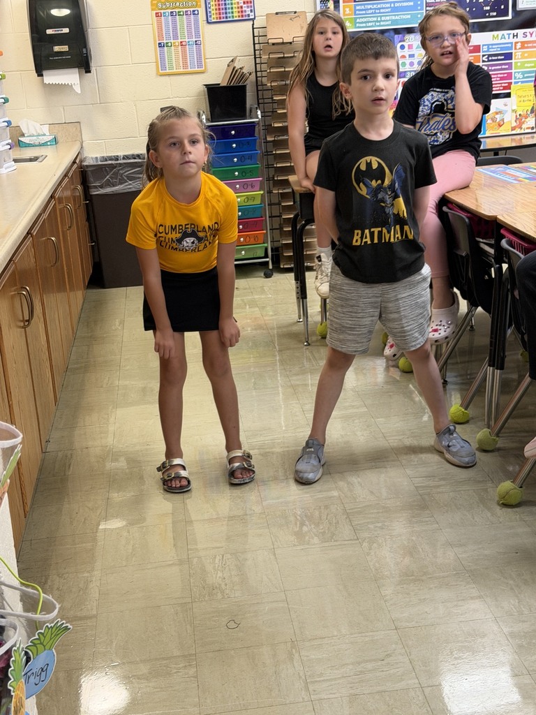 Two students are standing side by side answering a multiplication problem as fast as they can. Other students in the class are watching. 