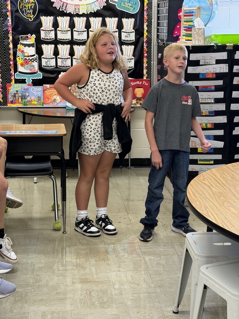 Two students are standing side by side answering a multiplication problem as fast as they can. Other students in the class are watching. 