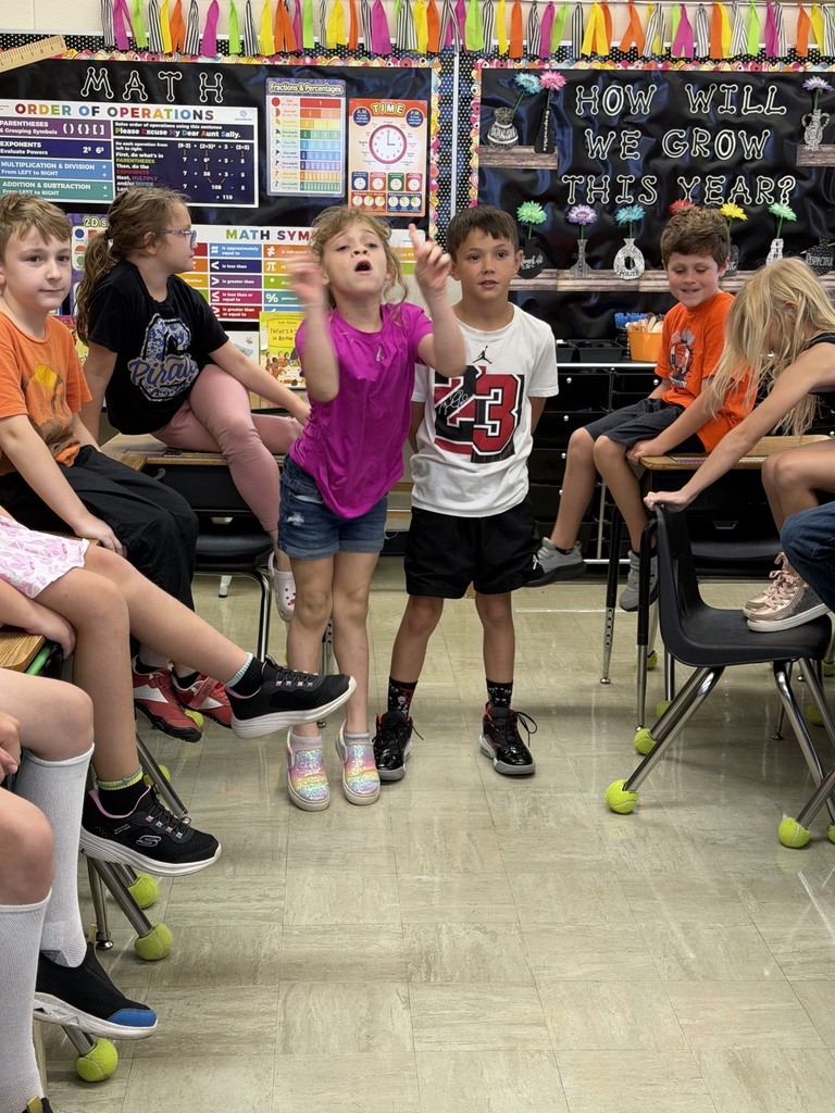 Two students are standing side by side answering a multiplication problem as fast as they can. Other students in the class are watching. 