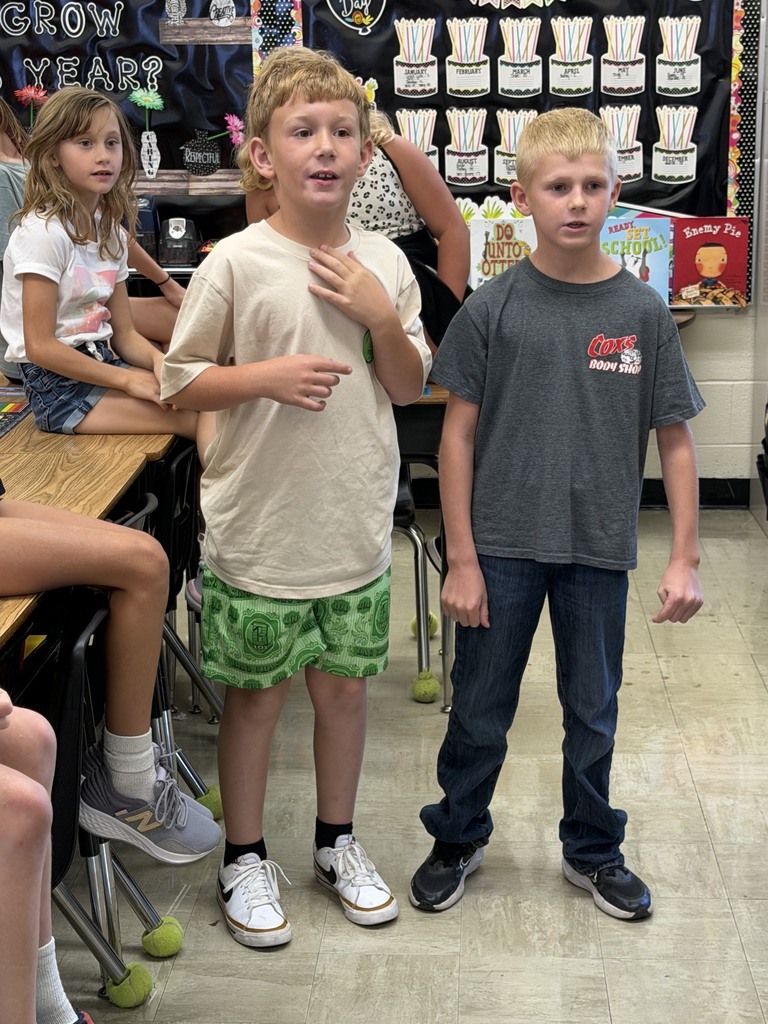 Two students are standing side by side answering a multiplication problem as fast as they can. Other students in the class are watching. 