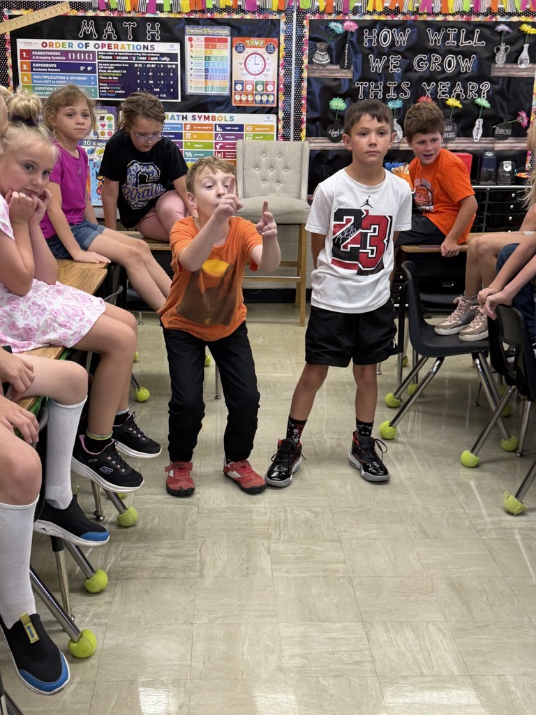 Two students are standing side by side answering a multiplication problem as fast as they can. Other students in the class are watching. 