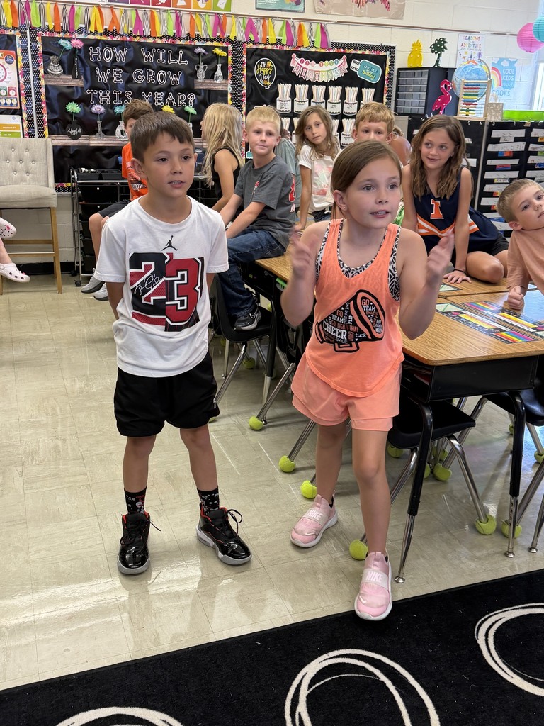 Two students are standing side by side answering a multiplication problem as fast as they can. Other students in the class are watching. 