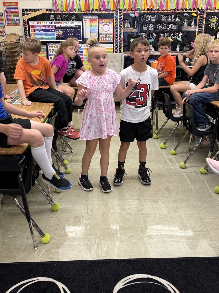 Two students are standing side by side answering a multiplication problem as fast as they can. Other students in the class are watching. 