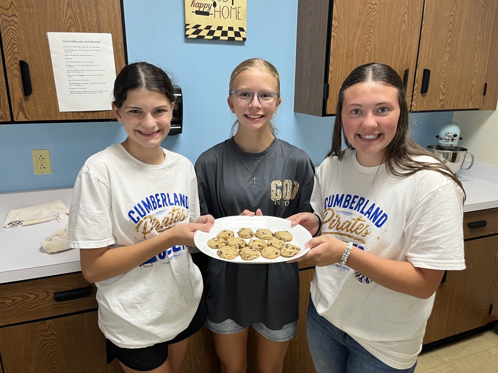Three students are standing in the kitchen. They are holding a platter of cookies. 
