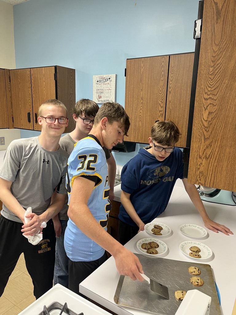 Four students are in the kitchen. One is removing cookies from a cookie sheet with a spatula. The others are watching. 