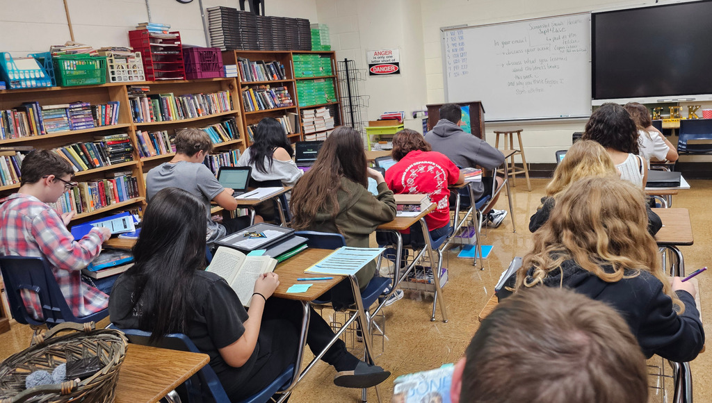 The students are facing away. They are sitting at their desks reading. 