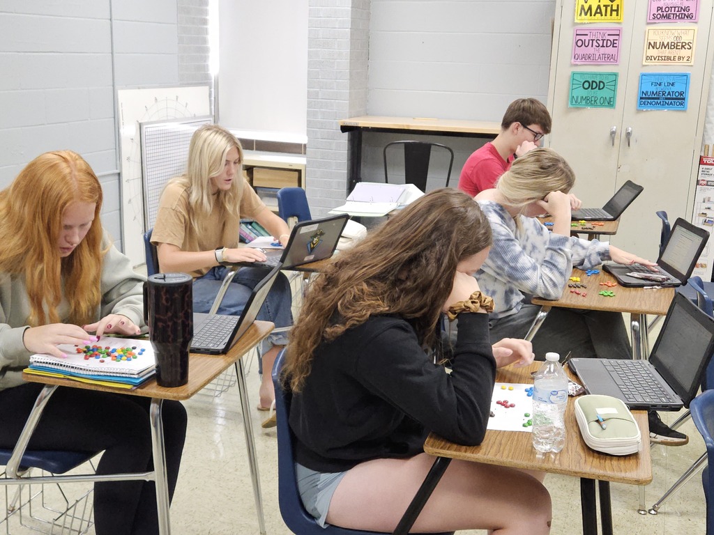 Students are sitting at desks. They have M&Ms spread out on their desks grouped by colors. They have open Chromebooks.