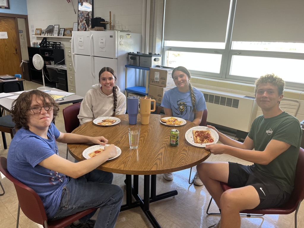 Four students are sitting at a table. There are four plates with pizza on each. 