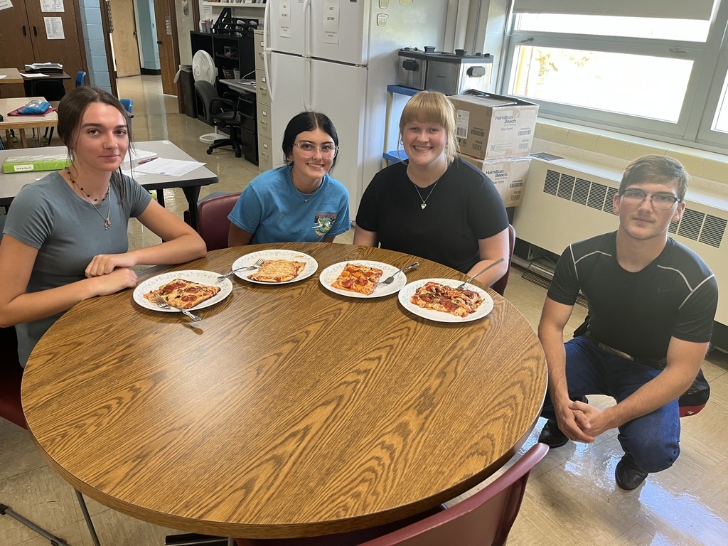 Four students are sitting at a table. There are four plates with pizza on each. 