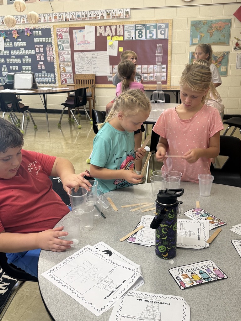Three students are standing behind a table. On the table are cups that the students are experimenting with. 