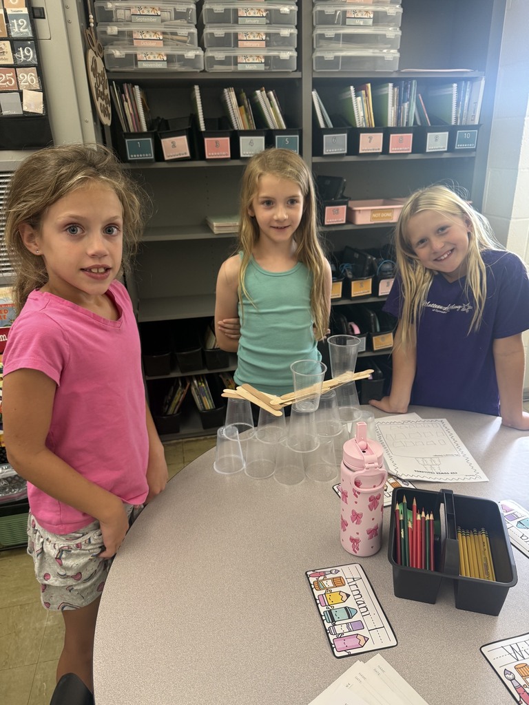 Three students are standing behind a table. On the table are cups stacked with popsicle sticks between them. 
