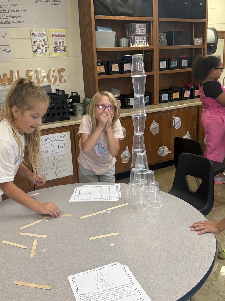 Two students are standing behind a table. One has their hands over their mouth in excitement. On the table are cups stacked with popsicle sticks between them. 