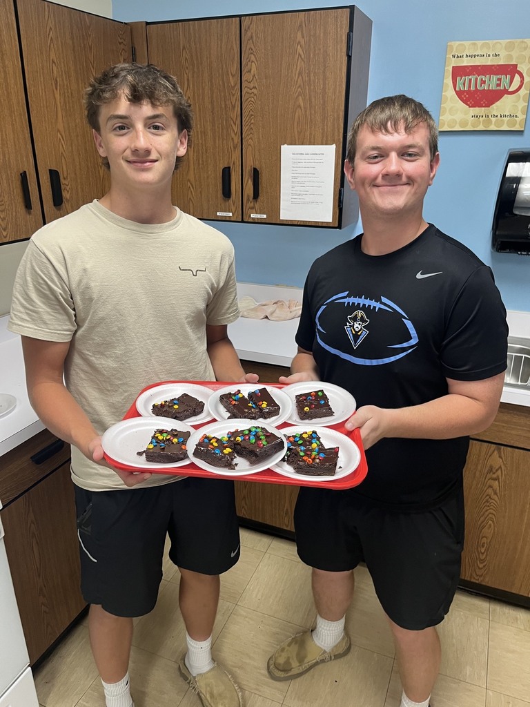 Two students are standing together. They are holding a tray with six plates of brownies.