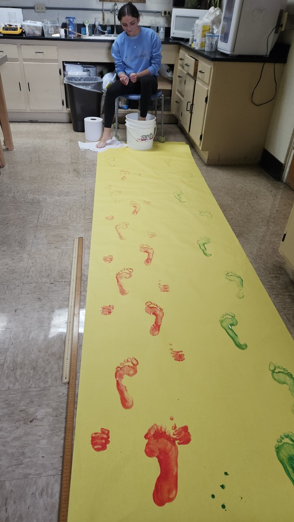 A student is sitting at the end of a long row of yellow paper. The student has their foot in a bucket cleaning it. There are painted foot prints on the yellow paper.