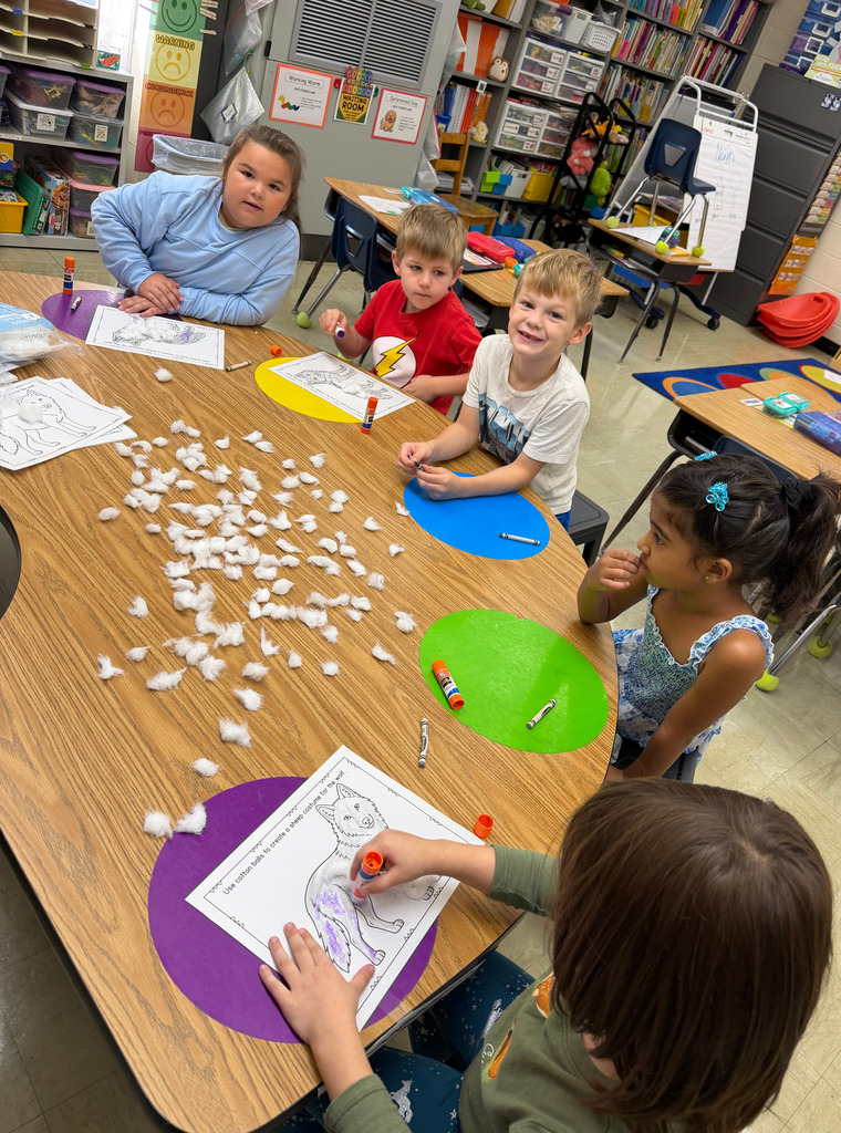 Students are sitting at a table. They are putting glue on their sheep. They are adding cotton to the glue.