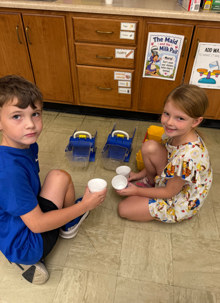 Two  students are sitting on the floor. They are holding white cups.They are looking at the camera.