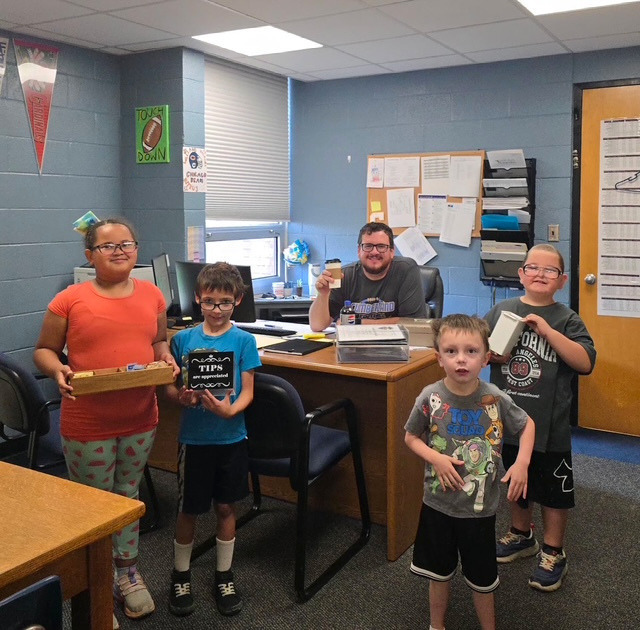 Four students are standing in front of the principal's desk holding a tip jar and condiments. The pricipal is holding up a cup of coffee.