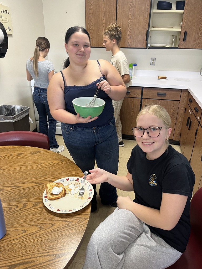 One student is sitting at a table with a plate of French toast. Another student has a bowl and a whisk.