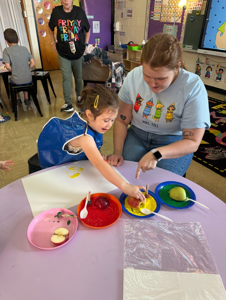 A student is dipping an apple in paint. The teacher is helping.