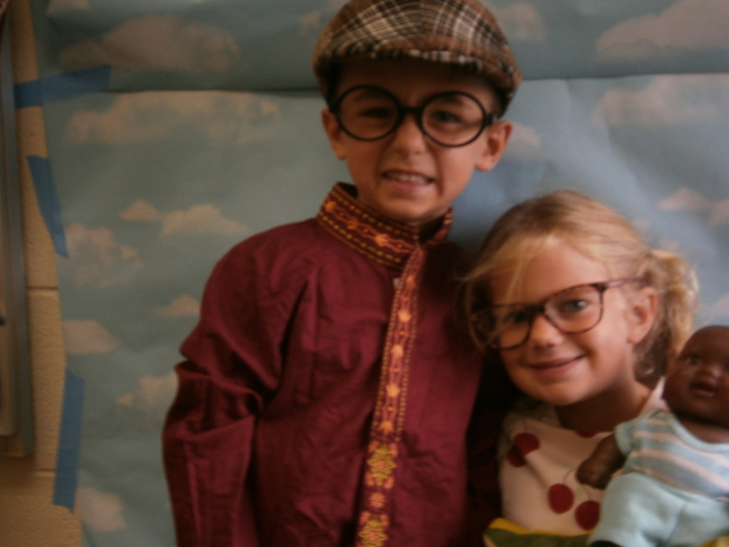 Two students are posing for a picture. Both have glasses. One has a hat and the other has a baby doll.