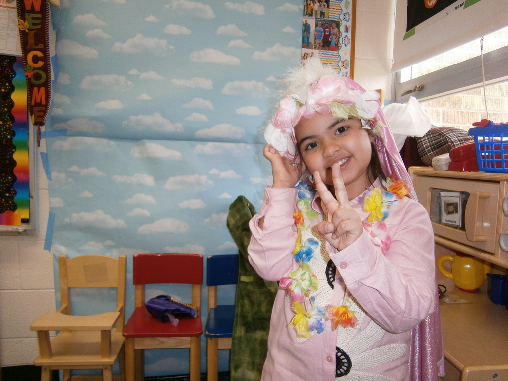 A student is wearing a cape, lei, and floral hat. The student is giving the peace sign.