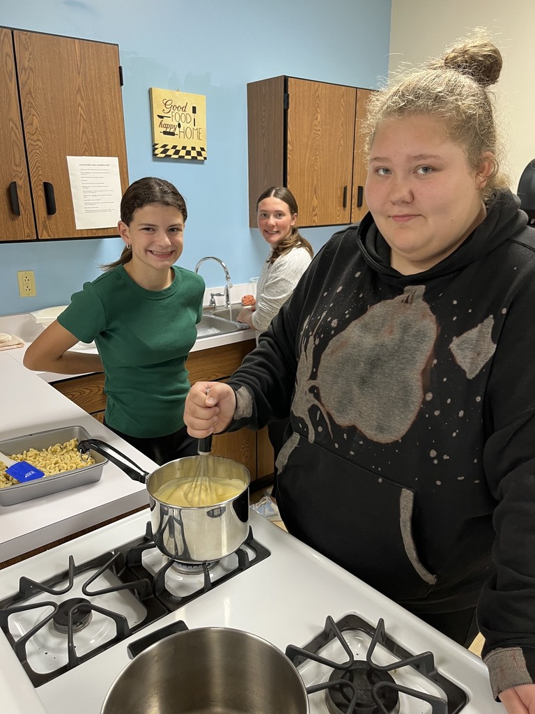 Three students are standing in their kitchen. One is stiring the cheese another is washing a dish.