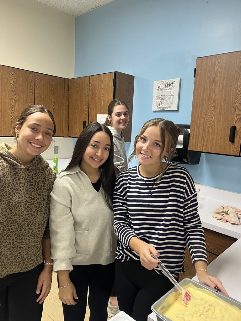 Three students are standing by a pan of mac and cheese. A fourth student is standing in the background.